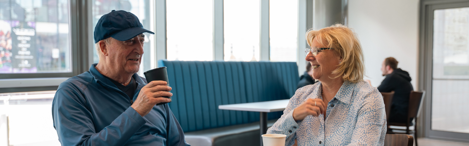 An older man and woman smiling and chatting together over coffee in a bright seating area.