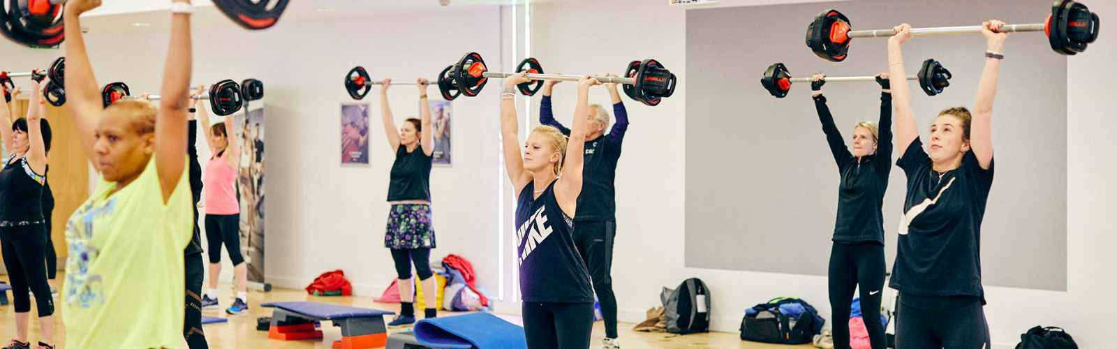Diverse group of participants performing overhead barbell presses in a lively group fitness class, promoting strength training and community wellness.