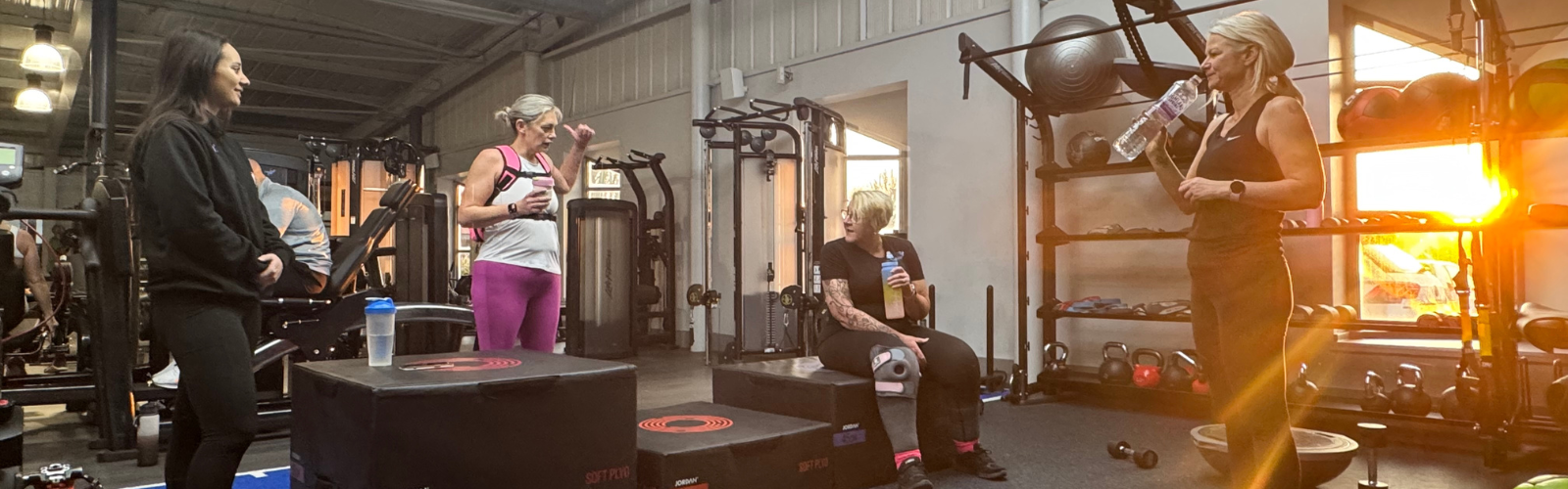 A group of women in a sunlit gym taking a break to chat and drink water after a workout.