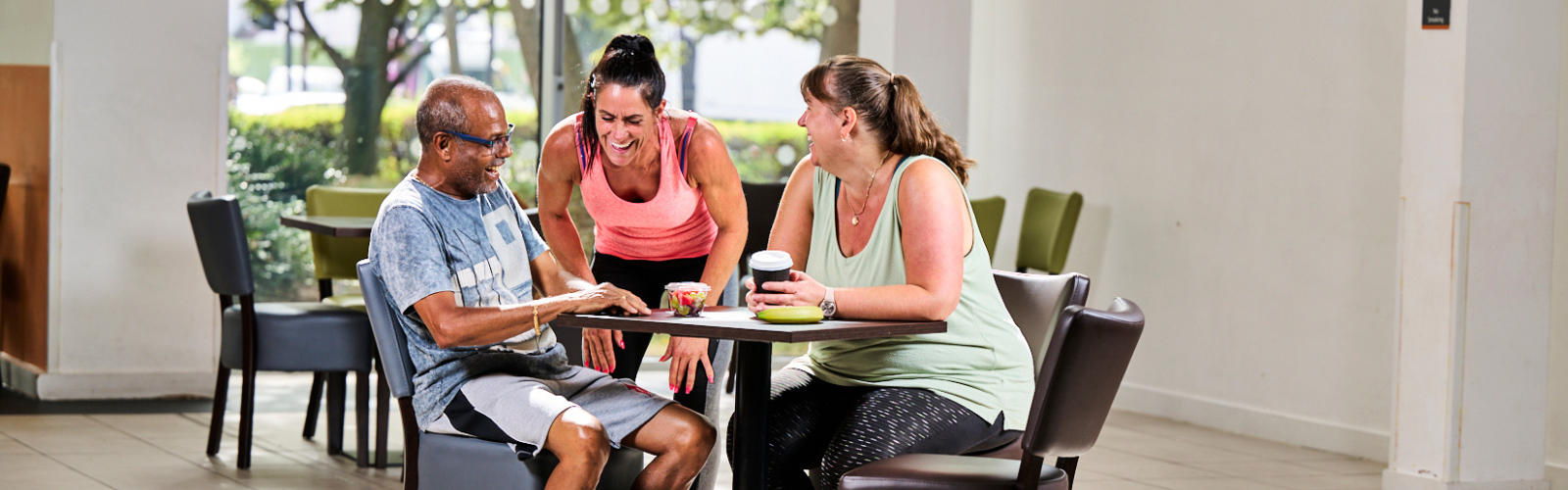 A fitness instructor and two members in athletic wear laughing together at a café table after a workout.