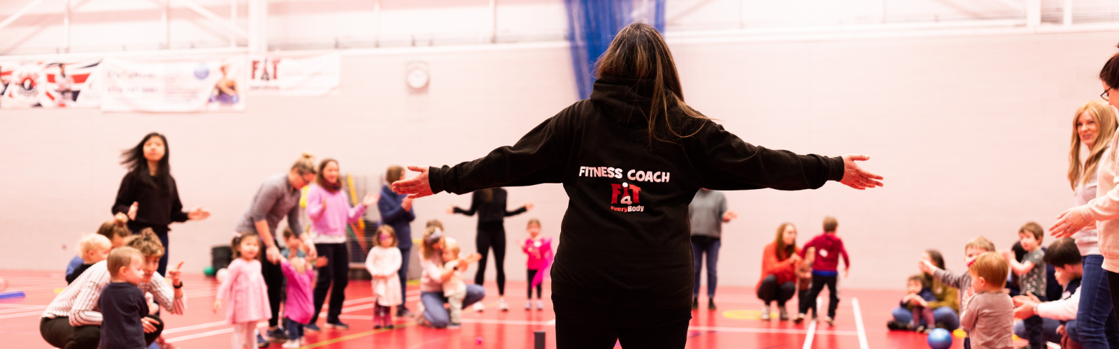 The back of a fitness coach with arms outstretched, leading a fun and active class for a large group of parents and young children in a sports hall.