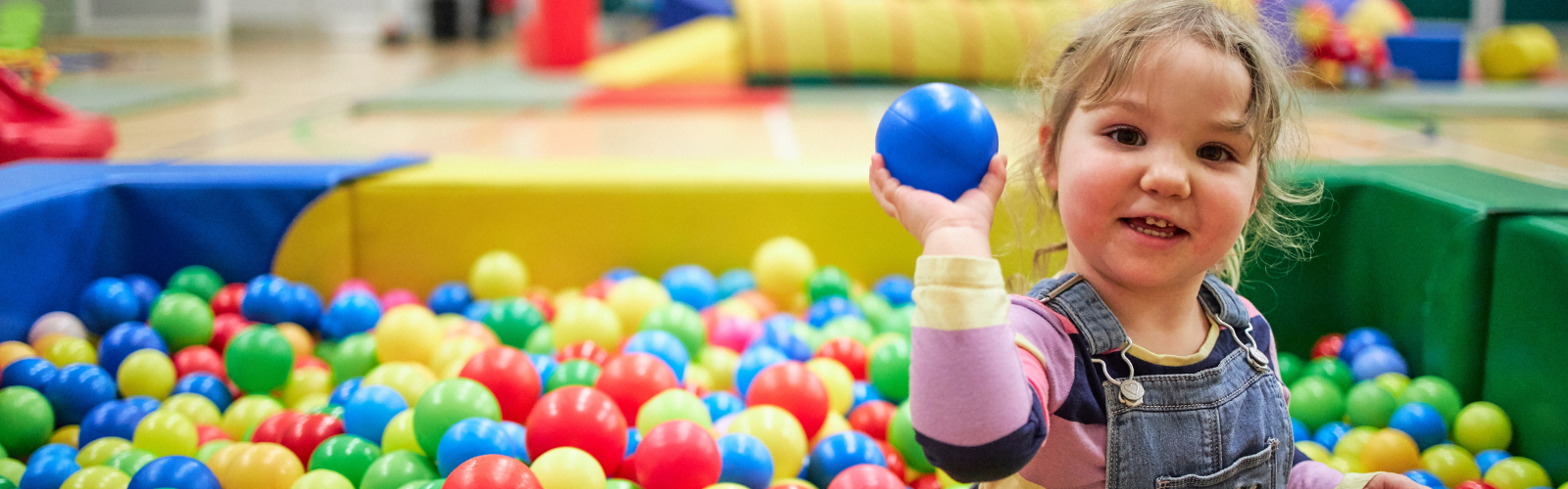 A young girl smiles at the camera while holding up a blue ball in a colourful soft-play ball pit.